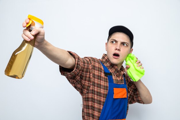 Young cleaning guy wearing uniform and cap holding rag with cleaning agent isolated on white wall