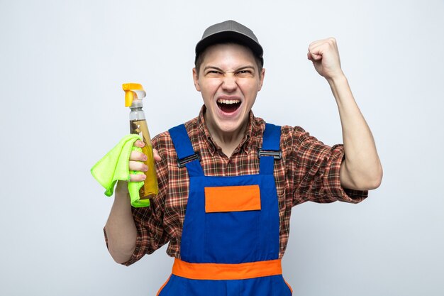 Young cleaning guy wearing uniform and cap holding rag with cleaning agent isolated on white wall