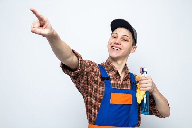 Young cleaning guy wearing uniform and cap holding rag with cleaning agent isolated on white wall