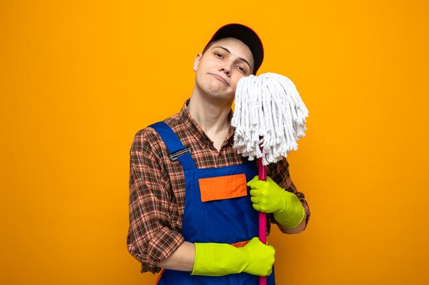 Young cleaning guy wearing uniform and cap with gloves holding mop isolated on orange wall