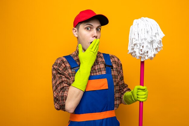 Young cleaning guy wearing uniform and cap with gloves holding mop isolated on orange wall