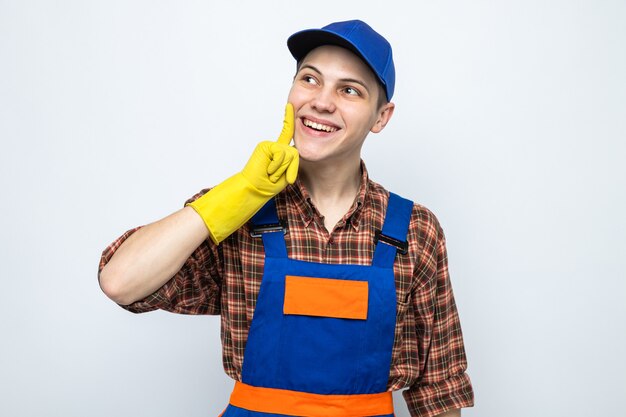 Young cleaning guy wearing uniform and cap with gloves isolated on white wall