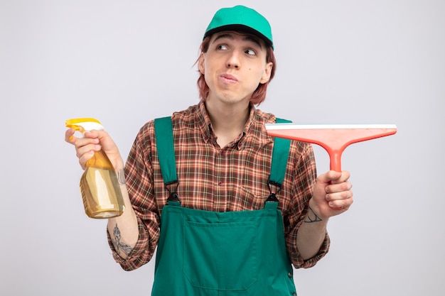 Young cleaning man in plaid shirt jumpsuit and cap holding cleaning spray and mop looking aside smiling confused standing on white