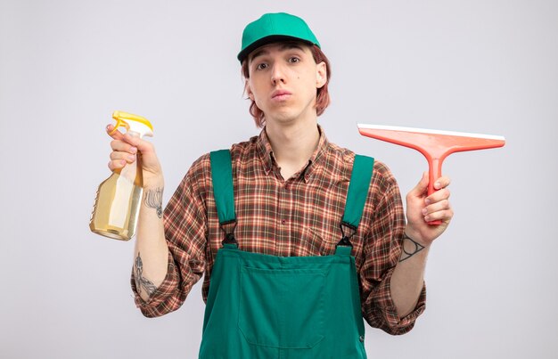 Young cleaning man in plaid shirt jumpsuit and cap holding cleaning spray and mop  with serious face standing over white wall