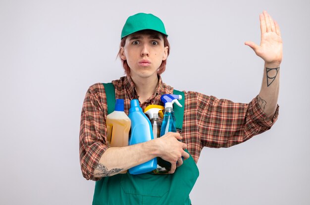 Young cleaning man in plaid shirt jumpsuit and cap holding cleaning supplies  worried raising hand standing over white wall