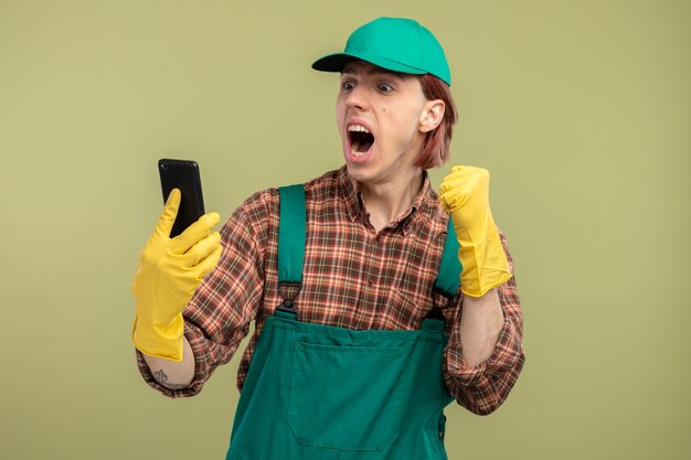 Young cleaning man in plaid shirt jumpsuit and cap wearing rubber gloves looking at his mobile phone happy and excited clenching fist