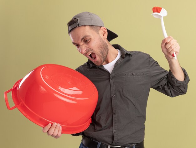 Free photo young cleaning man wearing casual clothes and cap holding basin and cleaning brush shouting with aggressive expression standing over green wall