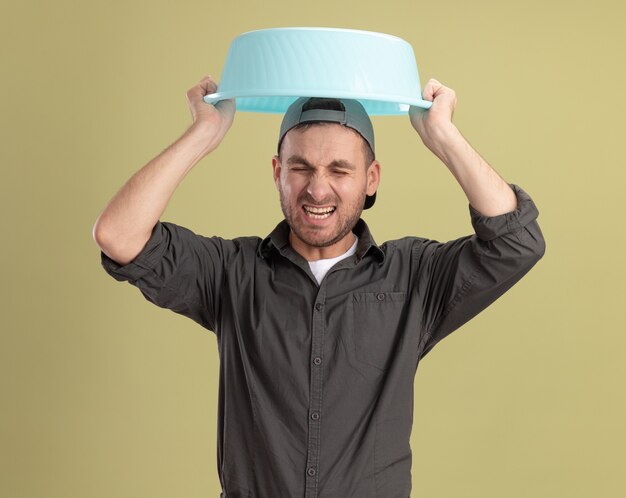 Young cleaning man wearing casual clothes and cap holding basin over her head with annoyed expression yelling standing over green wall