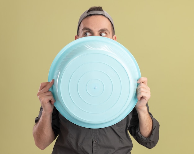 Free photo young cleaning man wearing casual clothes and cap holding basin hiding face behind it peeking over standing over green wall