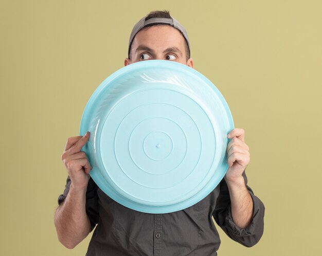 Young cleaning man wearing casual clothes and cap holding basin hiding face behind it peeking over standing over green wall