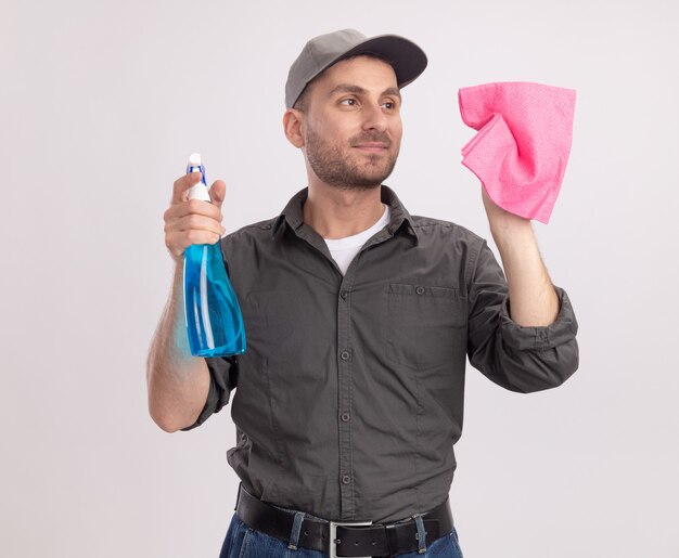 Young cleaning man wearing casual clothes and cap holding cleaning spray and rag ready to clean smiling confident standing over white wall