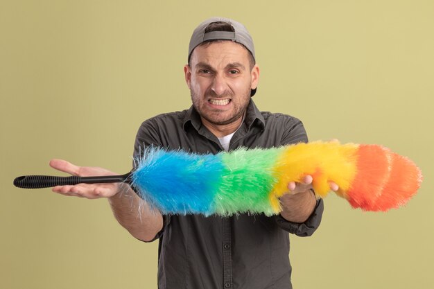 Young cleaning man wearing casual clothes and cap holding colorful duster looking  with annoyed expression displeased standing over green wall