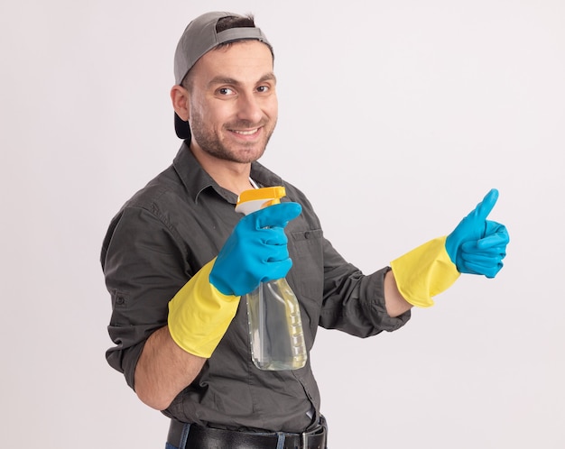 Молодой уборщик в повседневной одежде и кепке в резиновых перчатках держит чистящий спрей, улыбаясь, показывает палец вверх, стоя над оранжевой стеной