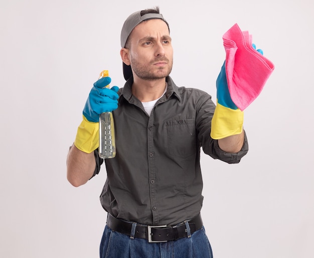 Молодой уборщик в повседневной одежде и кепке в резиновых перчатках держит чистящий спрей и тряпку, внимательно смотрит в сторону с серьезным лицом, стоящим над белой стеной