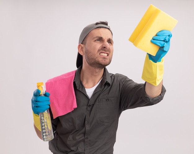 Young cleaning man wearing casual clothes and cap in rubber gloves holding cleaning spray and sponge with rag on his shoulder looking at sponge with annoyed expression standing over orange wall