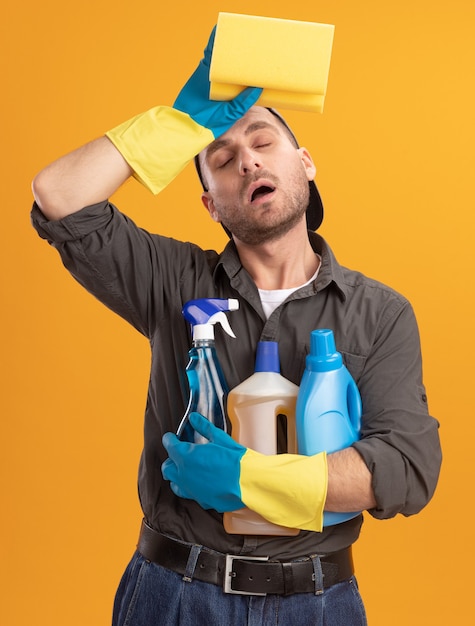 Foto gratuita giovane uomo delle pulizie che indossa abiti casual e berretto in guanti di gomma che tengono i prodotti per la pulizia e la spugna che sembra stanco e oberato di lavoro con la mano sulla fronte in piedi sopra la parete arancione