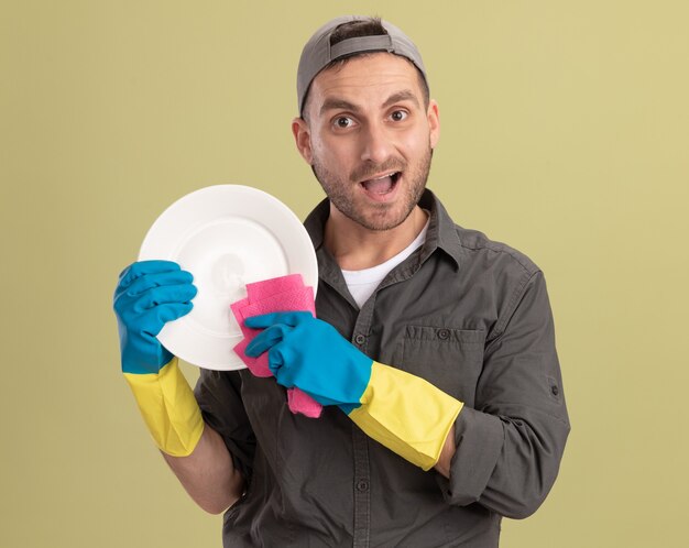 Young cleaning man wearing casual clothes and cap in rubber gloves holding plate and rag looking  happy and excited standing over green wall