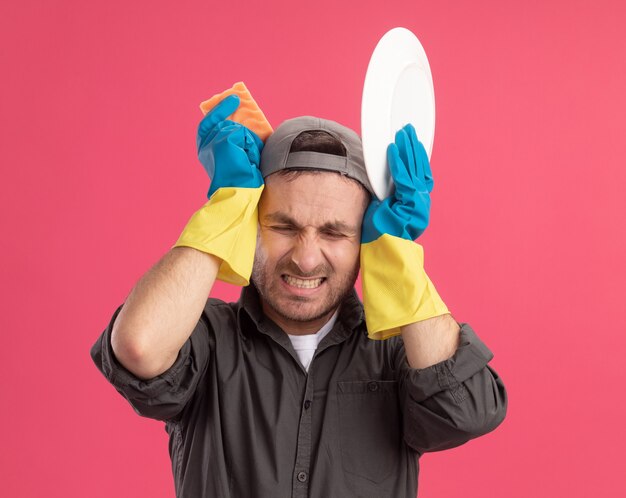 Молодой уборщик в повседневной одежде и кепке в резиновых перчатках держит тарелку и губку, закрывая уши с раздраженным выражением лица, стоит над розовой стеной