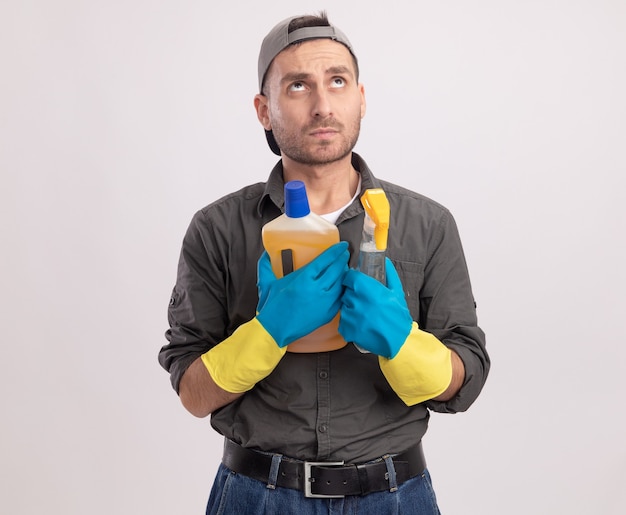 Young cleaning man wearing casual clothes and cap in rubber gloves holding spray bottle and cleaning supplies looking up puzzled standing over white wall