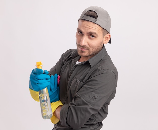 Giovane uomo delle pulizie che indossa abiti casual e berretto in guanti di gomma tenendo la bottiglia spray guardando con espressione scettica in piedi sopra il muro bianco