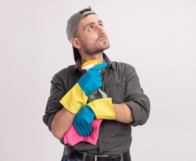 Young cleaning man wearing casual clothes and cap in rubber gloves holding spray bottle and rag looking aside puzzled standing over white wall