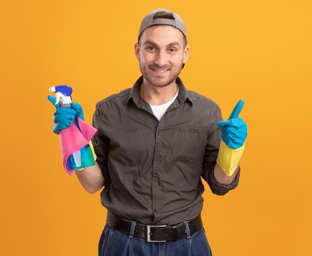 Young cleaning man wearing casual clothes and cap in rubber gloves holding spray bottle and rag looking  smiling cheerfully showign thumbs up standing over orange wall