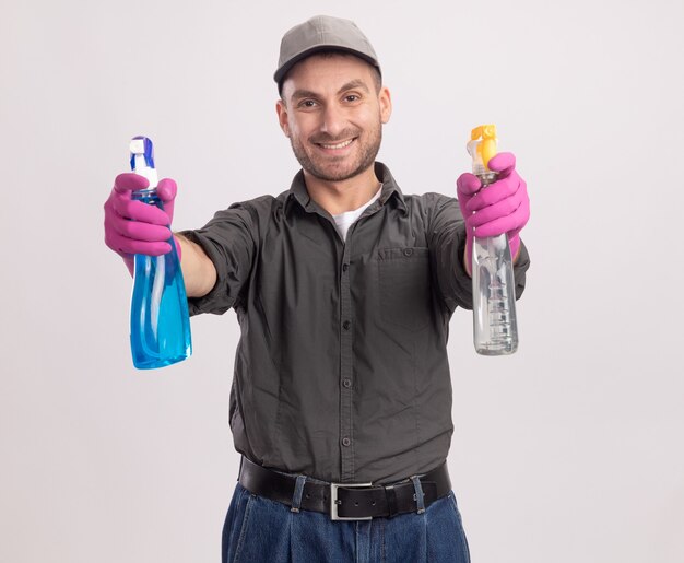 白い壁の上に元気に立って笑顔に見えるスプレーボトルを保持しているゴム手袋でカジュアルな服とキャップを身に着けている若い掃除人