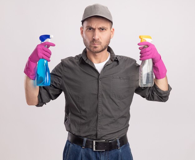 白い壁の上に立っている真面目な顔で見ているスプレーボトルを保持しているゴム手袋でカジュアルな服とキャップを身に着けている若い掃除人