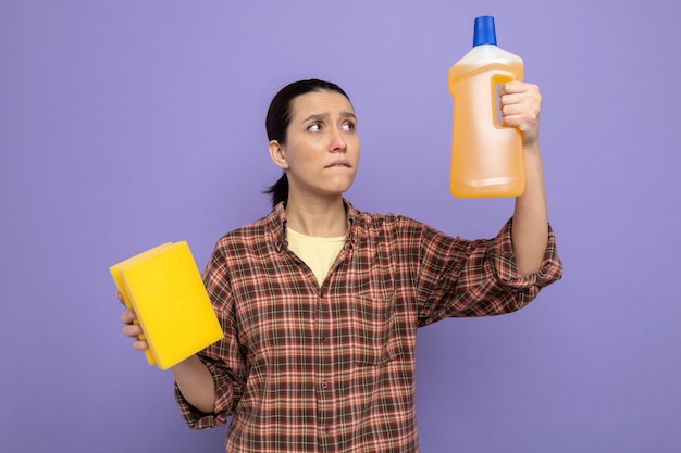 Young cleaning woman in casual clothes holding bottle of cleaning supplies with sponge looking confused trying to make choice standing on purple