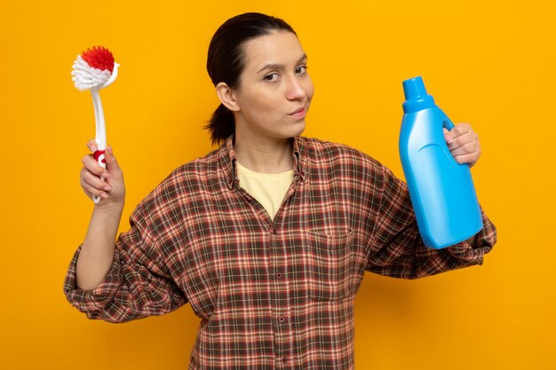 Young cleaning woman in casual clothes holding cleaning brush and bottle of cleaning supplies looking with serious face