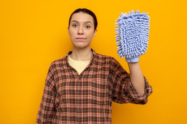 Young cleaning woman in casual clothes holding duster  with confident expression standing over orange wall