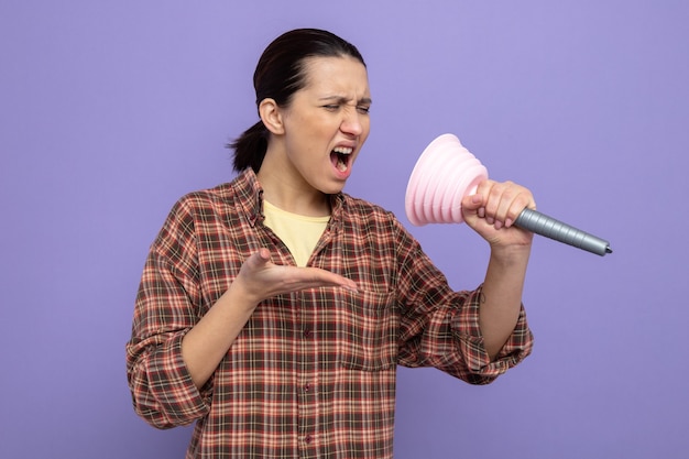 Young cleaning woman in casual clothes holding plunger using it as microphone singing having fun at work standing on purple