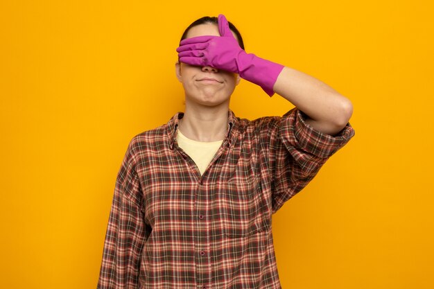 Young cleaning woman in casual clothes in rubber gloves coning eyes with hand standing on orange