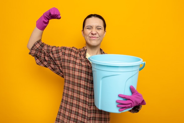 Young cleaning woman in casual clothes in rubber gloves holding bucket happy and excited clenching fist standing on orange