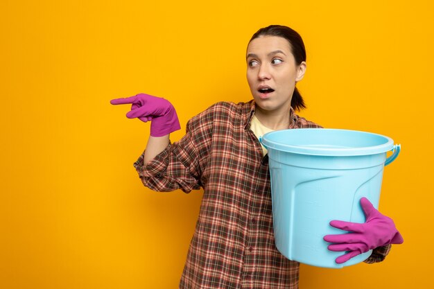 Foto gratuita giovane donna delle pulizie in abiti casual in guanti di gomma che tiene il secchio da parte preoccupato che punta con il dito indice sul lato in piedi sull'arancia