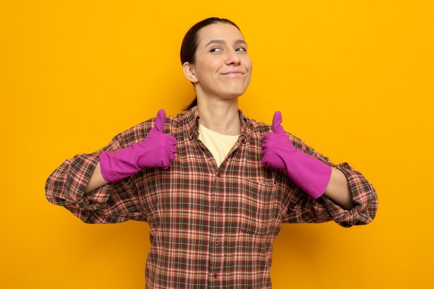 Young cleaning woman in casual clothes in rubber gloves looking aside with smile on happy face showing thumbs up standing over orange wall