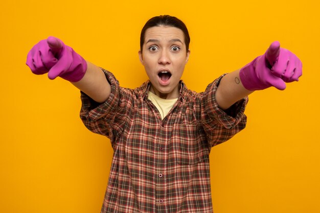 Young cleaning woman in casual clothes in rubber gloves looking surprised pointing with index fingers at front standing over orange wall