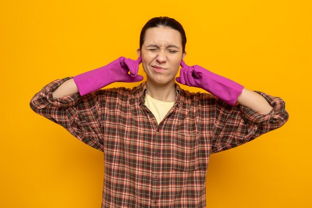 Young cleaning woman in casual clothes in rubber gloves looking unwell closing ears with fingers with annoyed expression standing over orange wall