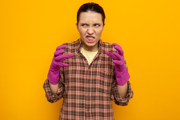 Молодая уборщица в повседневной одежде в резиновых перчатках заставляет скривленный рот злиться и разочаровываться поднятыми руками, стоя на оранжевом Бесплатные Фотографии