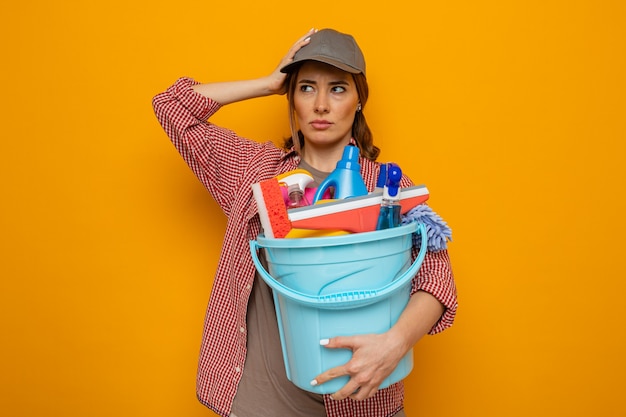 Young cleaning woman in plaid shirt and cap holding bucket with cleaning tools looking aside confused with hand on her head standing over orange background