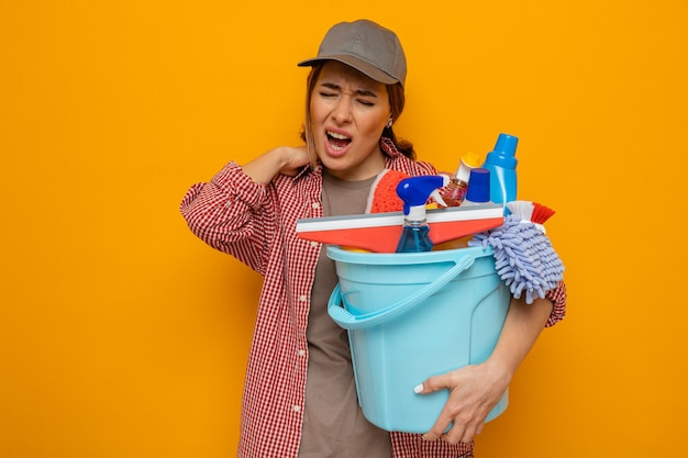 Young cleaning woman in plaid shirt and cap holding bucket with cleaning tools looking unwell touching her neck feeling pain standing over orange background