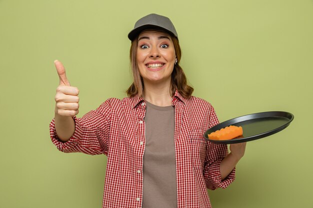 Young cleaning woman in plaid shirt and cap holding plate and sponge looking smiling showing thumbs up