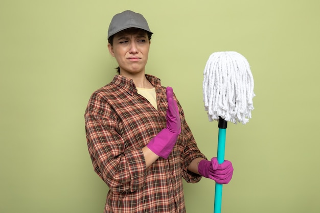 Young cleaning woman in plaid shirt and cap in rubber gloves holding mop looking at it with disgusted expression making defense gesture standing on green