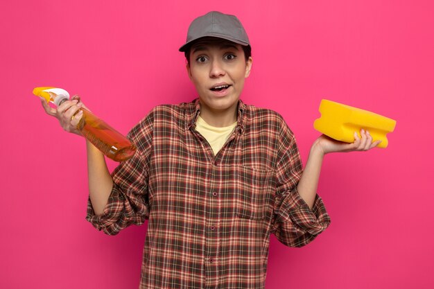 Young cleaning woman in plaid shirt and cap in rubber gloves holding sponge and cleaning spray confused spreading arms to the sides standing on pink