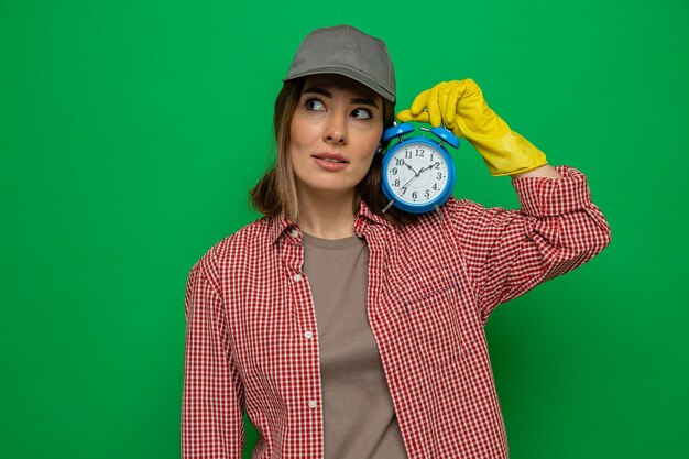 체크 무늬 셔츠와 모자를 쓴 젊은 청소부, 고무 장갑을 끼고 알람 시계를 들고 혼란스러운 모습