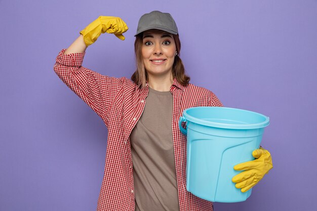 Free photo young cleaning woman in plaid shirt and cap wearing rubber gloves holding bucket looking smiling raising fist like a winner