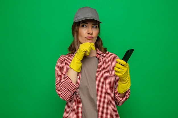 Giovane donna delle pulizie in camicia a quadri e berretto che indossa guanti di gomma che tengono lo smartphone guardando perplesso in piedi su sfondo verde