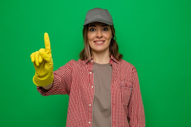 Молодая уборщица в клетчатой рубашке и кепке в резиновых перчатках смотрит в камеру, счастливая и удивленная, показывая указательный палец, имеющий новую идею, стоящий на зеленом фоне