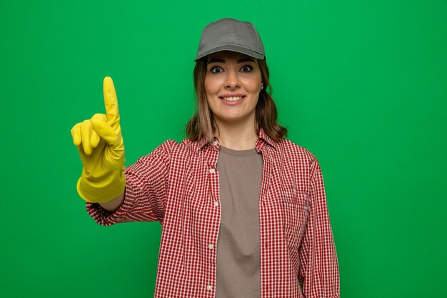 Young cleaning woman in plaid shirt and cap wearing rubber gloves looking at camera happy and surprised showing index finger having new idea standing over green background