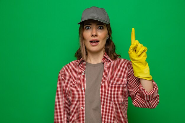 Free photo young cleaning woman in plaid shirt and cap wearing rubber gloves looking at camera surprised showing index finger having new idea standing over green background